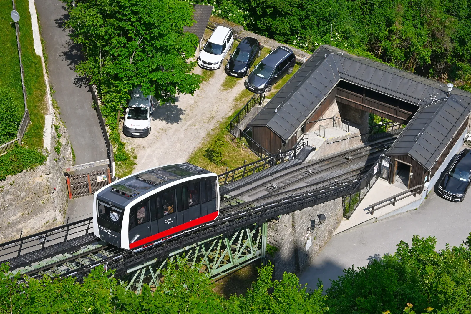 Seen from above, a funicular car at the lower station of the FestungsBahn among trees, with the station roof and a small car park beside it