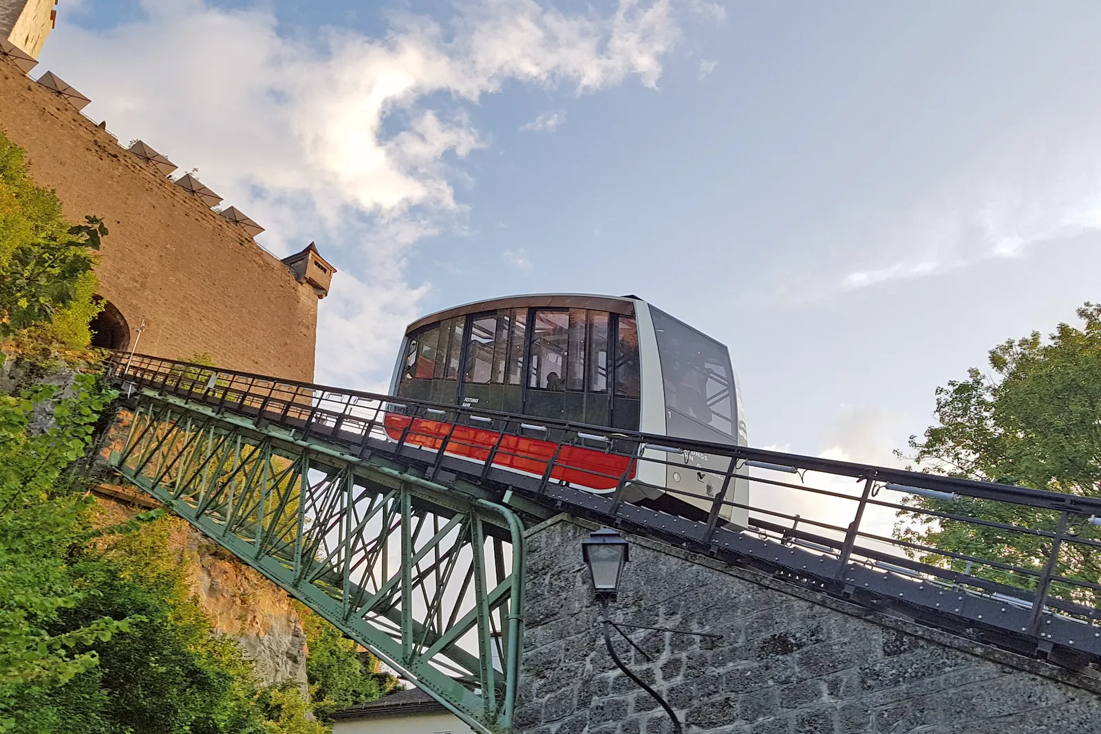 The FestungsBahn funicular car crossing a green steel bridge on its way up the hill, with a castle wall behind it and a blue sky above