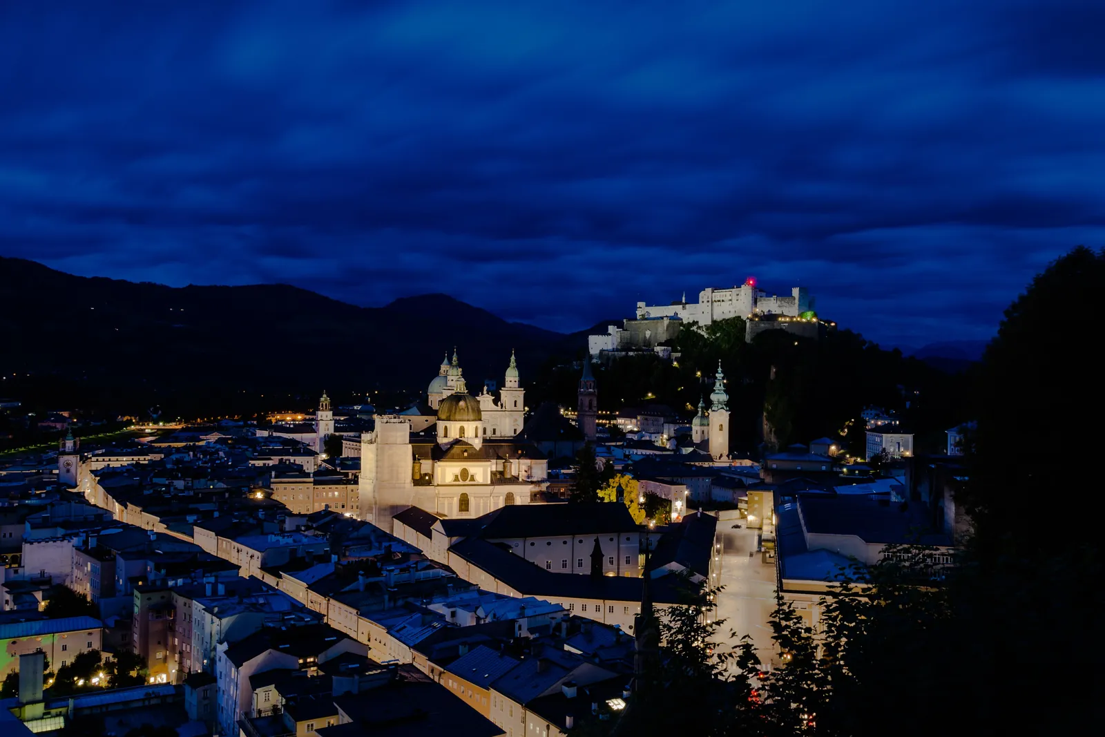 Salzburg's Old Town at dusk with lit streets and church domes, and the fortress glowing on the hill in the distance under a dark blue sky
